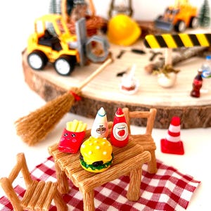 Pu&ograve; includere: Scena di picnic in miniatura con una tovaglia a scacchi rossi e bianchi, tavolo e sedie in legno e cibi come hamburger, patatine fritte e condimenti. Veicoli da costruzione e una scopa sullo sfondo.