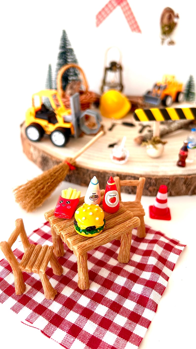 Pu&ograve; includere: Scena di picnic in miniatura con una tovaglia a scacchi rossi e bianchi, tavolo e sedie in legno e cibi come hamburger, patatine fritte e condimenti. Veicoli da costruzione e una scopa sullo sfondo.