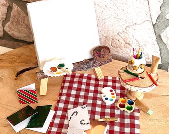 Può includere: Un atelier d'artista in miniatura con un cavalletto di legno, una tela bianca, palette di colori, pennelli e un tavolino con una tovaglia a quadri rossi e bianchi.