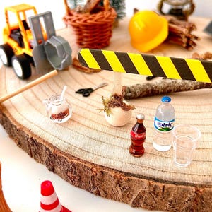 Pu&ograve; includere: Scena di cantiere in miniatura su una fetta di legno. Include un escavatore giocattolo giallo e grigio, un cestino in miniatura, un casco giallo, una bottiglia di Coca-Cola, una bottiglia d'acqua e coni stradali rossi e bianchi. Sono presenti anche un alberello e una lanterna.
