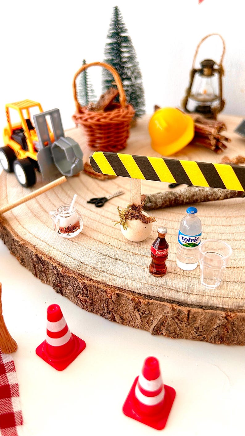 Pu&ograve; includere: Scena di cantiere in miniatura su una fetta di legno. Include un escavatore giocattolo giallo e grigio, un cestino in miniatura, un casco giallo, una bottiglia di Coca-Cola, una bottiglia d'acqua e coni stradali rossi e bianchi. Sono presenti anche un alberello e una lanterna.