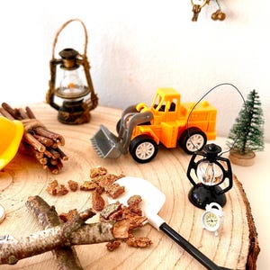 Pu&ograve; includere: Una scena in miniatura con una pala caricatrice giocattolo gialla, una lanterna nera e una piccola sveglia bianca. Sono presenti anche un albero di Natale in miniatura e un fascio di ramoscelli. La scena &egrave; ambientata su una fetta di legno.