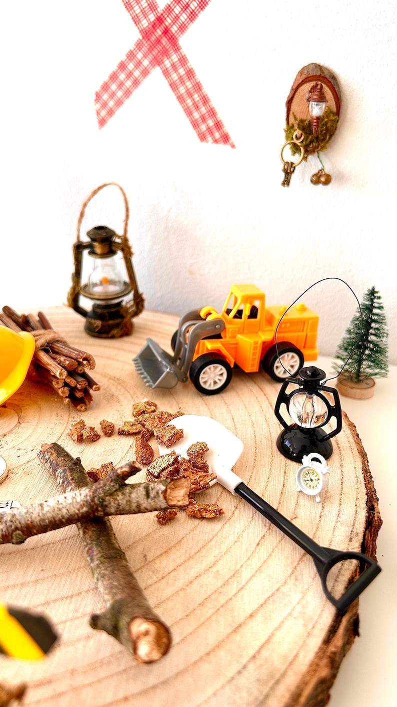 Pu&ograve; includere: Una scena in miniatura con una pala caricatrice giocattolo gialla, una lanterna nera e una piccola sveglia bianca. Sono presenti anche un albero di Natale in miniatura e un fascio di ramoscelli. La scena &egrave; ambientata su una fetta di legno.