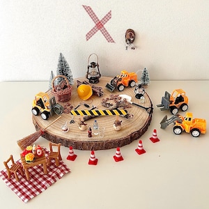 Pu&ograve; includere: Una scena di cantiere in miniatura con camion giocattolo, coni e un tavolo da picnic su una fetta di legno. La scena comprende un casco giallo, una scopa e un cestino. Una coperta da picnic a scacchi rossi e bianchi &egrave; stesa.