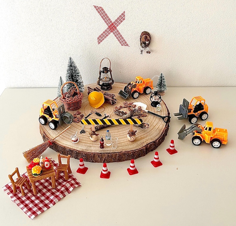 Pu&ograve; includere: Una scena di cantiere in miniatura con camion giocattolo, coni e un tavolo da picnic su una fetta di legno. La scena comprende un casco giallo, una scopa e un cestino. Una coperta da picnic a scacchi rossi e bianchi &egrave; stesa.