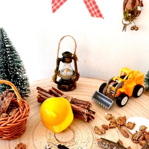 Pu&ograve; includere: Una scena di costruzione in miniatura su una superficie di legno. Include un casco giallo, un bulldozer giocattolo, una lanterna, un cestino e degli attrezzi. Elementi decorativi: nastro a scacchi rosso e bianco e un piccolo albero di Natale.