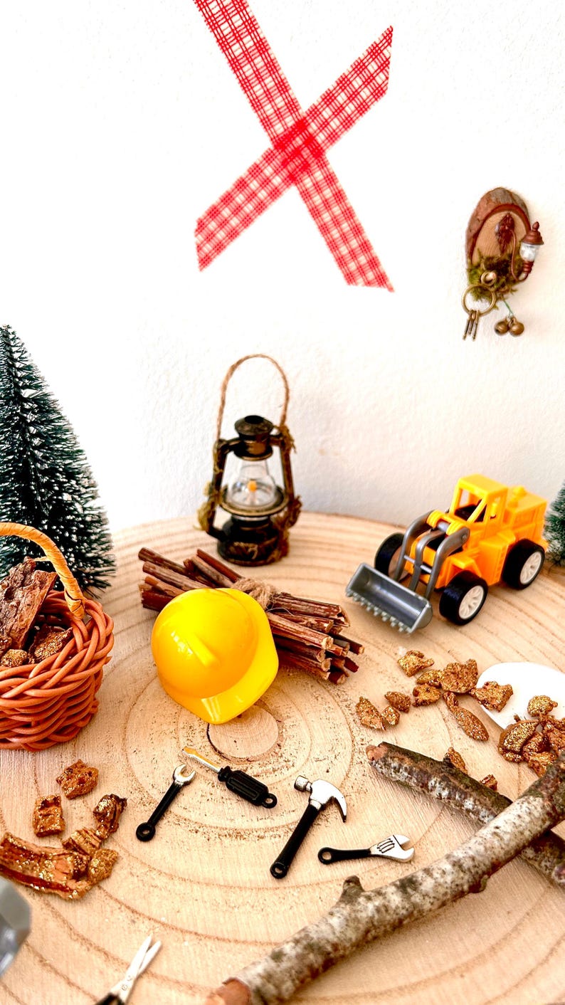Pu&ograve; includere: Una scena di costruzione in miniatura su una superficie di legno. Include un casco giallo, un bulldozer giocattolo, una lanterna, un cestino e degli attrezzi. Elementi decorativi: nastro a scacchi rosso e bianco e un piccolo albero di Natale.