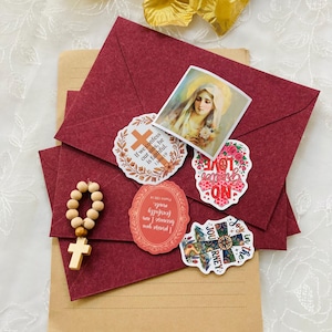 May include: A dark red envelope adorned with five religious stickers, including an image of the Virgin Mary, a cross, and text-based designs. A small wooden rosary with a cross charm rests on the left side of the envelope, which is placed atop a tan paper folder.