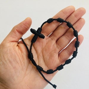 May include: Black knotted bracelet with a cross charm. The bracelet has small knots that resemble beads.