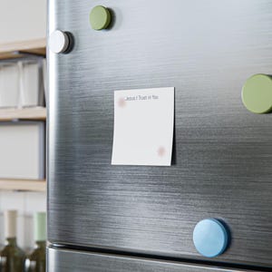 May include: A square white sticky note attached to a brushed stainless steel refrigerator. The note features the printed text Jesus I Trust in You in the upper left corner, accompanied by two small, light pink starburst or floral graphic icons.