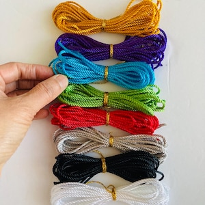 May include: A collection of colorful, tightly wound cords in shades of gold, purple, turquoise, green, red, gray, black, and white. Each cord is secured with a gold-colored tie. The cords are likely used for crafting or decorative purposes.
