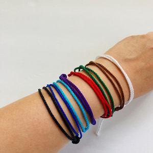 May include: A collection of colorful rope bracelets in various shades, including black, blue, purple, red, green, brown, and white. The bracelets are simple and minimalist, suitable for everyday wear. The bracelets are tied with a knot.