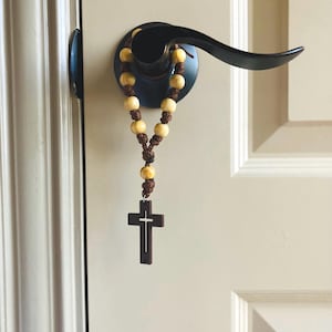 May include: A rosary featuring light tan wooden beads strung on a dark brown cord, ending in a dark brown wooden cross with a cutout center design. The rosary is looped around a black metal door handle attached to a light-colored paneled door.