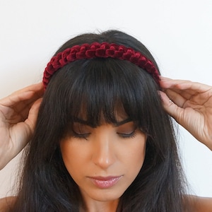 Puede incluir: Una diadema de terciopelo burdeos con un diseño texturizado y tejido. La diadema se lleva en la cabeza de una persona, con cabello oscuro que enmarca la cara. El fondo es blanco liso.