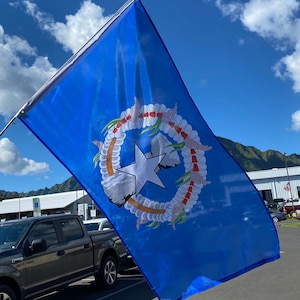 May include: A blue flag with a white star and a wreath of white flowers with red centers. The flag is flying in the wind against a blue sky with white clouds.