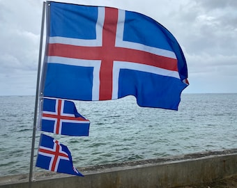 May include: Three Icelandic flags waving in the wind against a backdrop of blue ocean and a cloudy sky. The largest flag is blue with a white cross and a red cross on top of the white cross. The two smaller flags are the same design but smaller in size.