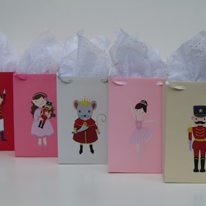 May include: Five gift bags with different Nutcracker-themed designs. The bags are red, pink, white, pink, and beige. Each bag has a different character from the Nutcracker story, including a soldier, a ballerina, a mouse king, a ballerina, and a nutcracker.