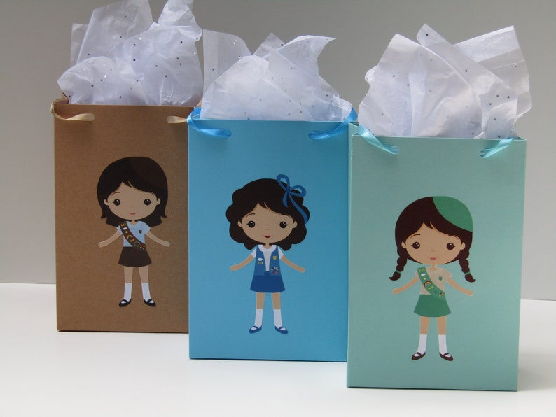 May include: Three gift bags with tissue paper inside. Each bag has a different color and features a cartoon girl scout wearing a different uniform. The bags are brown, blue, and green.