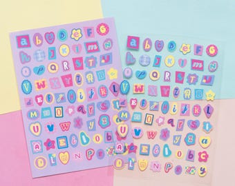 Könnte beinhalten: Zwei Bögen mit Alphabet-Aufklebern in verschiedenen Formen und Farben. Die Aufkleber zeigen Buchstaben A bis Z, Zahlen und Symbole in einer Pastellfarbpalette. Ein Bogen hat einen lila Hintergrund, der andere ist transparent.