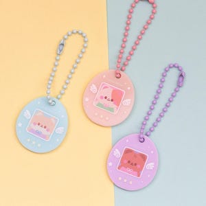 May include: Three pastel-coloured keychains with cartoon bear designs. The keychains are oval-shaped with a ball chain and a clasp. The colours are light blue, pink, and purple. Each keychain has a different bear design and three stars.