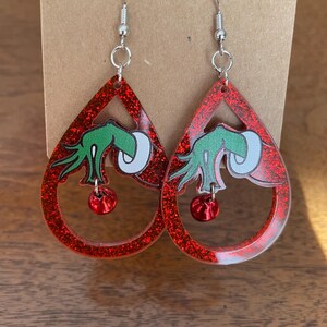 Puede incluir: Un par de pendientes rojos con purpurina en forma de lágrima. Cada pendiente presenta un gráfico verde y blanco y una pequeña cuenta roja. Los pendientes tienen ganchos para las orejas plateados.