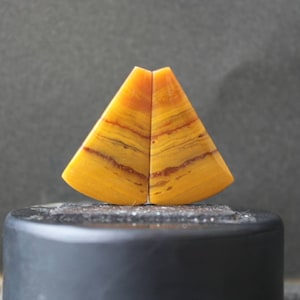 May include: Two yellow and orange agate cabochons with a striated pattern. The cabochons are shaped like a triangle with a rounded base.
