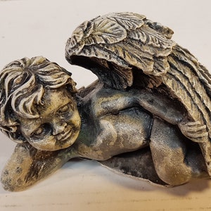 Peut inclure: Une petite figurine d'ange en pierre grise avec des ailes. L'ange est couch&eacute; sur le c&ocirc;t&eacute;, la t&ecirc;te reposant sur son bras. L'ange a une expression douce et innocente sur son visage.