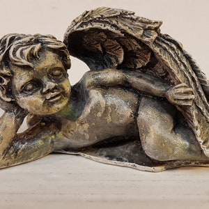 Peut inclure: Une petite figurine de ch&eacute;rubin ail&eacute; grise avec une finition vieillie. Le ch&eacute;rubin est couch&eacute; sur le c&ocirc;t&eacute;, la t&ecirc;te reposant sur sa main. Les ailes sont d&eacute;ploy&eacute;es derri&egrave;re le ch&eacute;rubin.