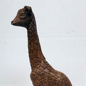 May include: A brown ceramic giraffe figurine with a textured surface, standing on a base with a rough texture.