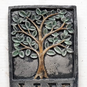 Op de afbeelding: Een grijze stenen plaquette met een reliëfbeeld van een boom met groene bladeren en een bruine stam. Het woord "LIFE" is onder de boom gegraveerd in gouden letters.