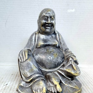 May include: A gray and gold painted ceramic statue of a smiling Buddha sitting in a meditative pose. The statue has a round base and is decorated with a textured pattern.