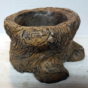 Puede incluir: Una maceta de cerámica marrón con forma de tronco de árbol con una abertura circular en la parte superior. La maceta tiene una superficie rugosa y texturizada que se asemeja a la corteza.
