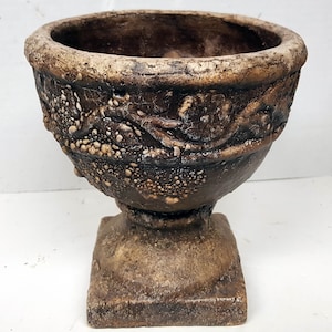 May include: A brown ceramic planter with a textured surface and a decorative design. The planter has a wide, round top and a square base.