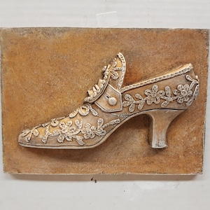 May include: A decorative wall plaque featuring a white ceramic shoe with floral details on a brown background.