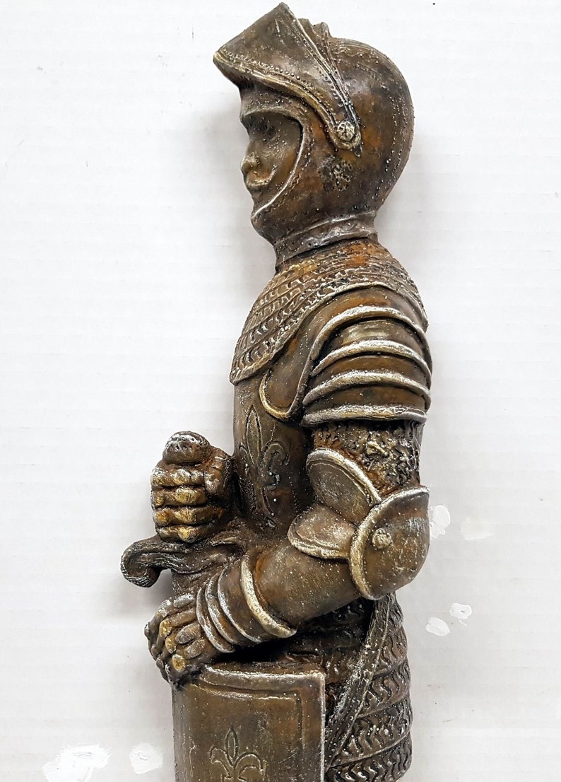 Puede incluir: Una figurita de caballero de color bronce con una espada y un escudo. El caballero lleva una armadura con un dise&ntilde;o de flor de lis.