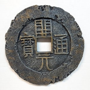 May include: A round, gray, metal coin with a square hole in the center. The coin has Chinese characters on it, including the character "元" (yuan).