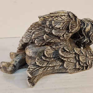 Peut inclure: Une petite figurine d'ange en pierre grise avec des ailes repli&eacute;es derri&egrave;re le dos. L'ange est couch&eacute; sur le c&ocirc;t&eacute;, la t&ecirc;te reposant sur ses ailes.