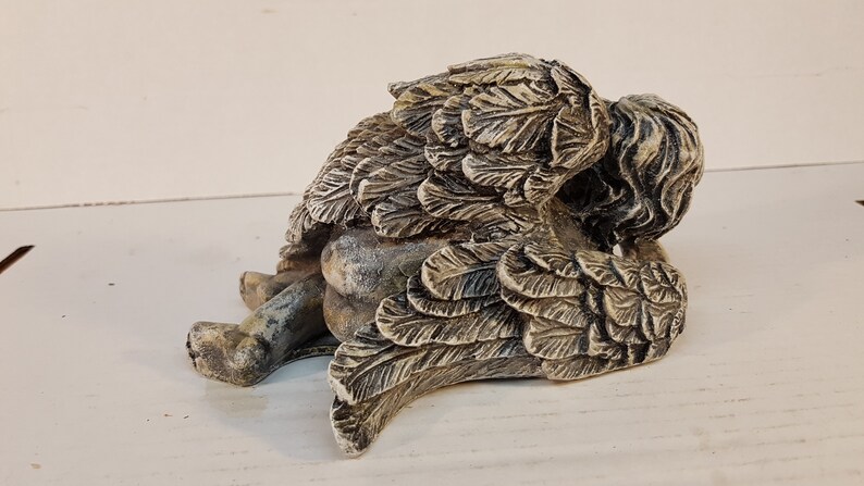 Peut inclure: Une petite figurine d'ange en pierre grise avec des ailes repli&eacute;es derri&egrave;re le dos. L'ange est couch&eacute; sur le c&ocirc;t&eacute;, la t&ecirc;te reposant sur ses ailes.