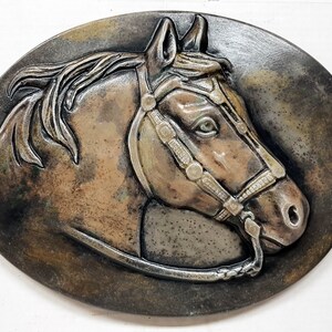 Può includere: Una scultura di testa di cavallo marrone con un morso in pelle, su uno sfondo marrone scuro testurizzato. La scultura è in ceramica e ha un aspetto realistico.