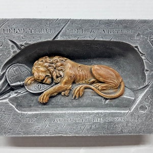 Puede incluir: Una placa de color plateado con una imagen en relieve de un león acostado sobre una roca. El león sostiene un escudo con una cruz. La placa tiene el texto "Helvetorum Fidei ac Virtuti" y "Die X Augusti MD.XCII" inscrito en ella.