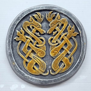 May include: A circular, gray stone plaque with a gold Celtic knot design featuring two intertwined dragons. The dragons are facing each other with their heads raised and their tails intertwined.