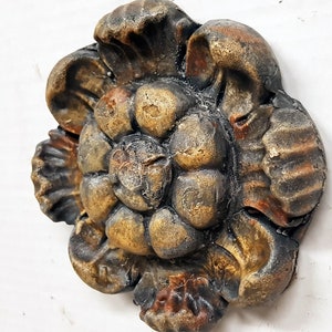 May include: A detailed, antique-style decorative floral element. The piece features a layered design with a mix of rounded and petal-like shapes. The color palette includes shades of gold, brown, and black, creating a weathered appearance. The object is likely made of a hard material.