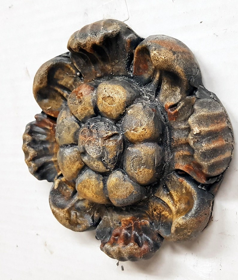 May include: A detailed, antique-style decorative floral element. The piece features a layered design with a mix of rounded and petal-like shapes. The color palette includes shades of gold, brown, and black, creating a weathered appearance. The object is likely made of a hard material.