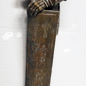 Puede incluir: Una estatua de metal marr&oacute;n y dorado de un caballero sosteniendo una espada. El caballero lleva una armadura con un dise&ntilde;o de flor de lis. La estatua est&aacute; montada sobre una base con una textura rugosa.