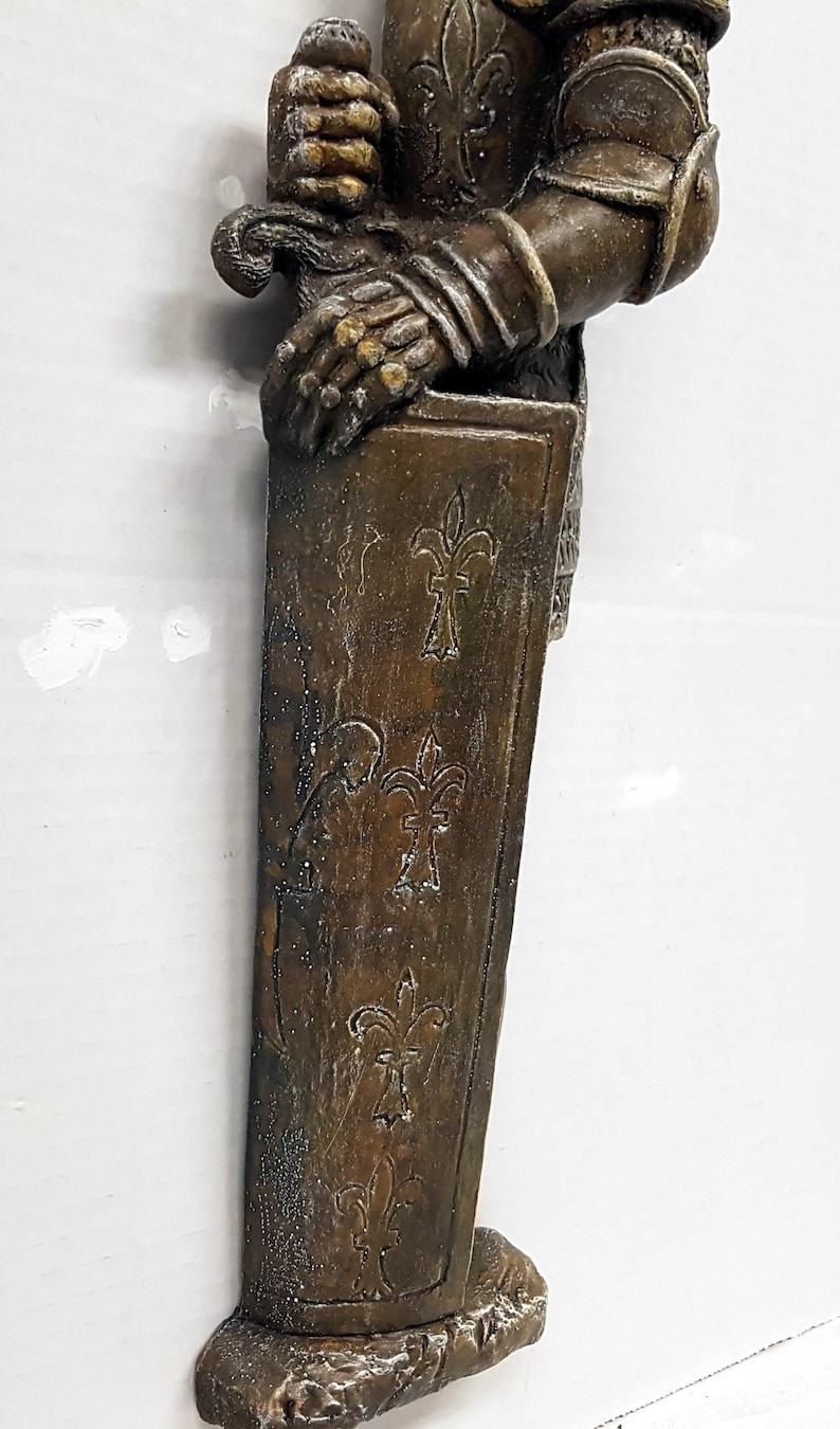 Puede incluir: Una estatua de metal marr&oacute;n y dorado de un caballero sosteniendo una espada. El caballero lleva una armadura con un dise&ntilde;o de flor de lis. La estatua est&aacute; montada sobre una base con una textura rugosa.