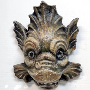 May include: A detailed, hand-painted, ceramic wall sculpture of a mythical creature with a fish-like head, large eyes, and a toothy grin. The sculpture is painted in shades of brown, green, and gray.