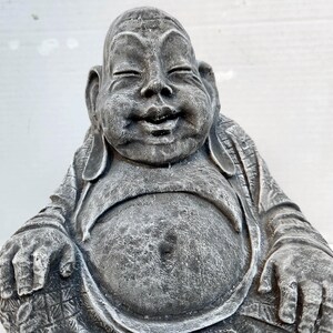 May include: A gray stone statue of a smiling Buddha sitting in a meditative pose. The statue has a round, full belly and is wearing a robe with intricate details.