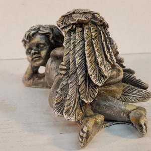 Peut inclure: Une paire de figurines de ch&eacute;rubins, l'une avec des ailes en plumes, l'autre avec une simple t&ecirc;te. Les ch&eacute;rubins sont faits d'une pierre gris clair avec des accents bruns et sont en position allong&eacute;e.