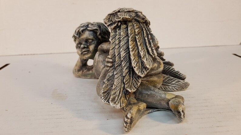 Peut inclure: Une paire de figurines de ch&eacute;rubins, l'une avec des ailes en plumes, l'autre avec une simple t&ecirc;te. Les ch&eacute;rubins sont faits d'une pierre gris clair avec des accents bruns et sont en position allong&eacute;e.