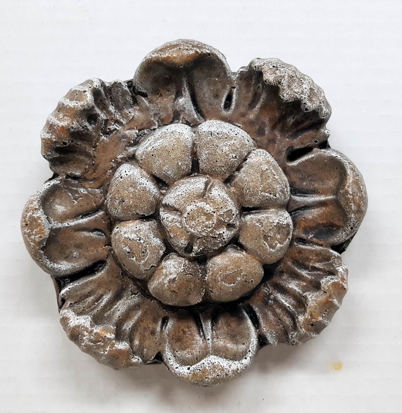 May include: A detailed, weathered, flower-shaped decorative element. The piece is primarily brown with white accents, giving it a textured, aged appearance. The design features layered petals and a central floral pattern.